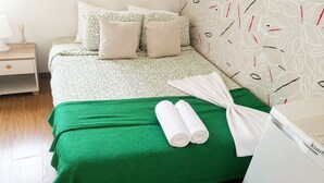 A neatly made bed with a green bedspread and two rolled white towels, a bedside table with a lamp, and a wall with a leaf pattern. - Room-charming Lisbon Apartment in Quiet (Lisbon)
