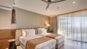 Een moderne hotelkamer met twee bedden, een plafondventilator, een groot raam met gordijnen, en een klein nachtkastje.