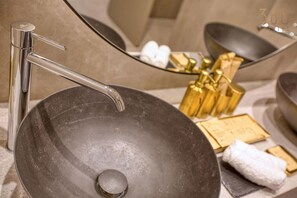 Ein modernes Badezimmer mit einem großen, runden Waschbecken, einem chromfarbenen Wasserhahn und goldfarbenen Pflegeprodukten.