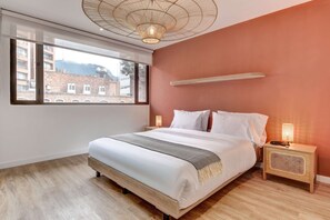 Ein Schlafzimmer mit einem großen Bett, einem Nachttisch mit Lampe, einem Fenster mit Blick auf Gebäude und einem Deckenventilator.
