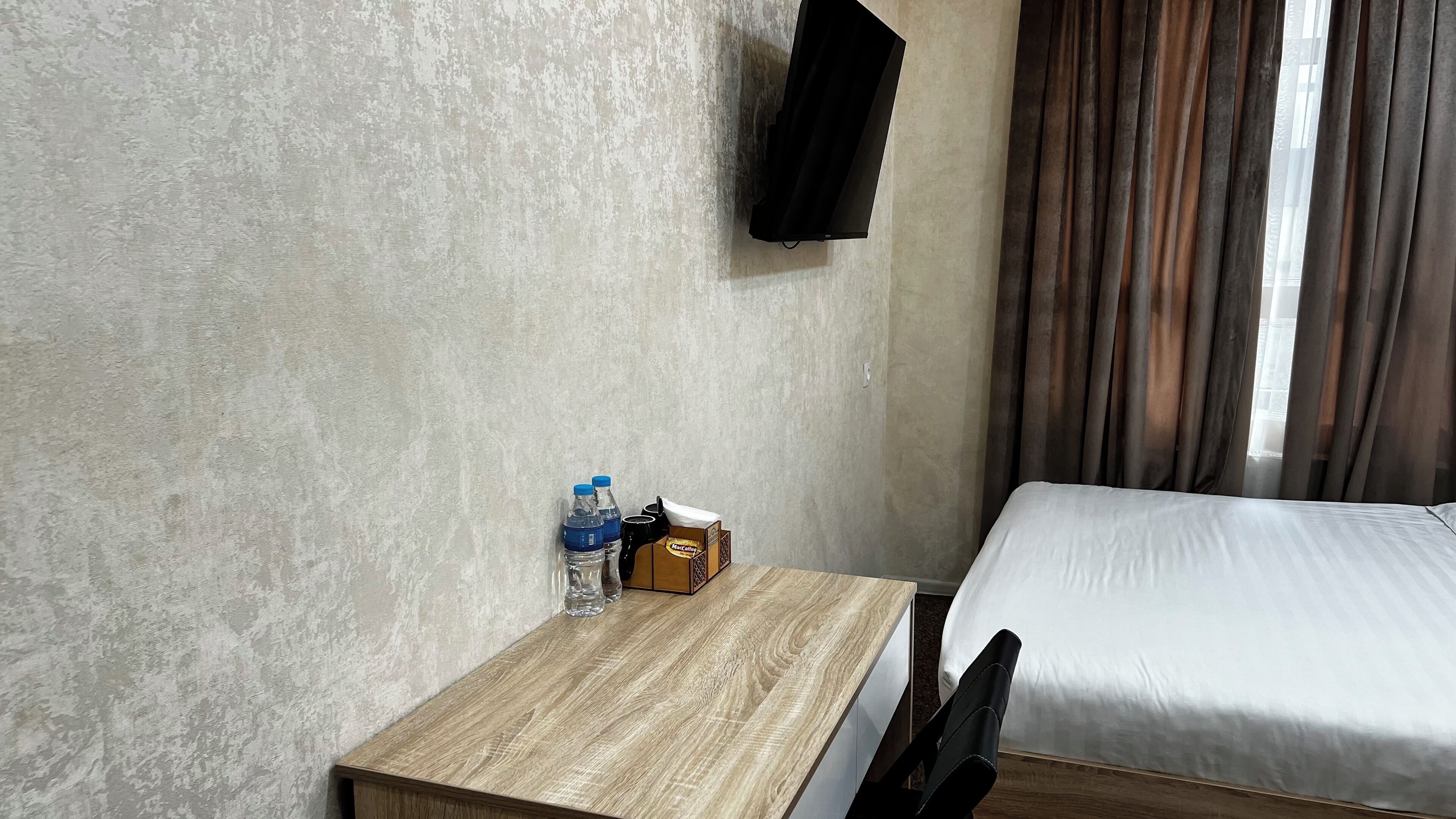A hotel room with a wooden desk, a bed, a flat-screen TV mounted on the wall, and a window with curtains.