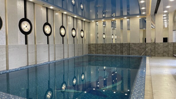A modern indoor swimming pool with a clear glass bottom, surrounded by walls with circular light fixtures and a tiled floor. - Chinor Hotel Fergana (Fergana)