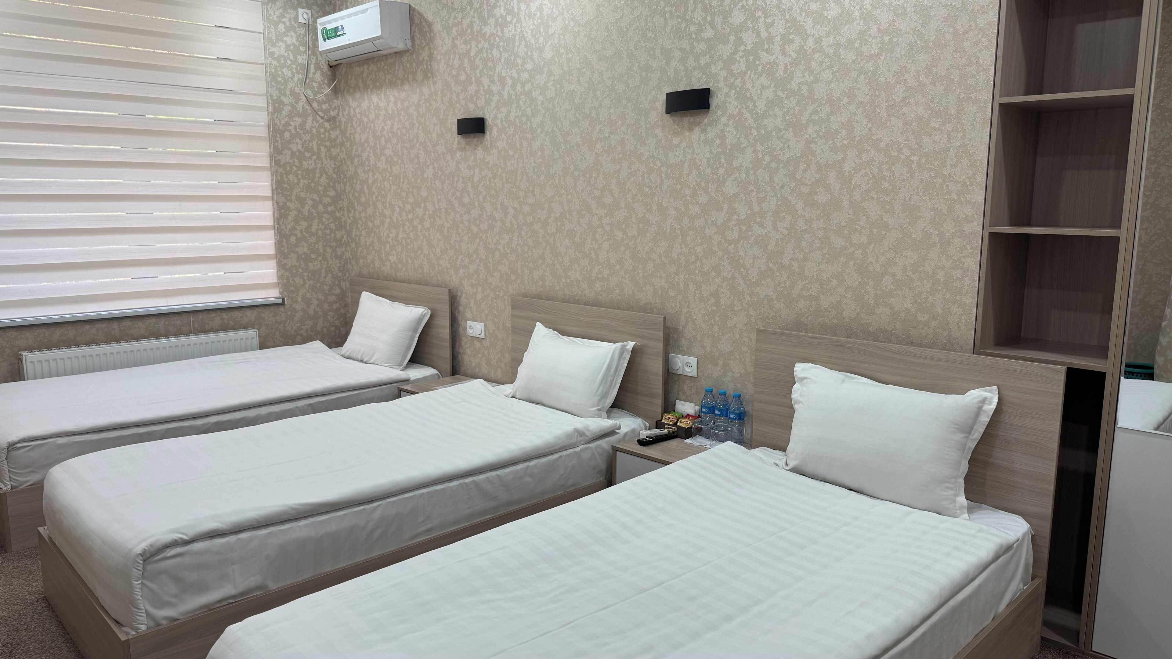 A hotel room with two single beds, a bedside table with a remote control, a water bottle, and a thermostat on the wall.