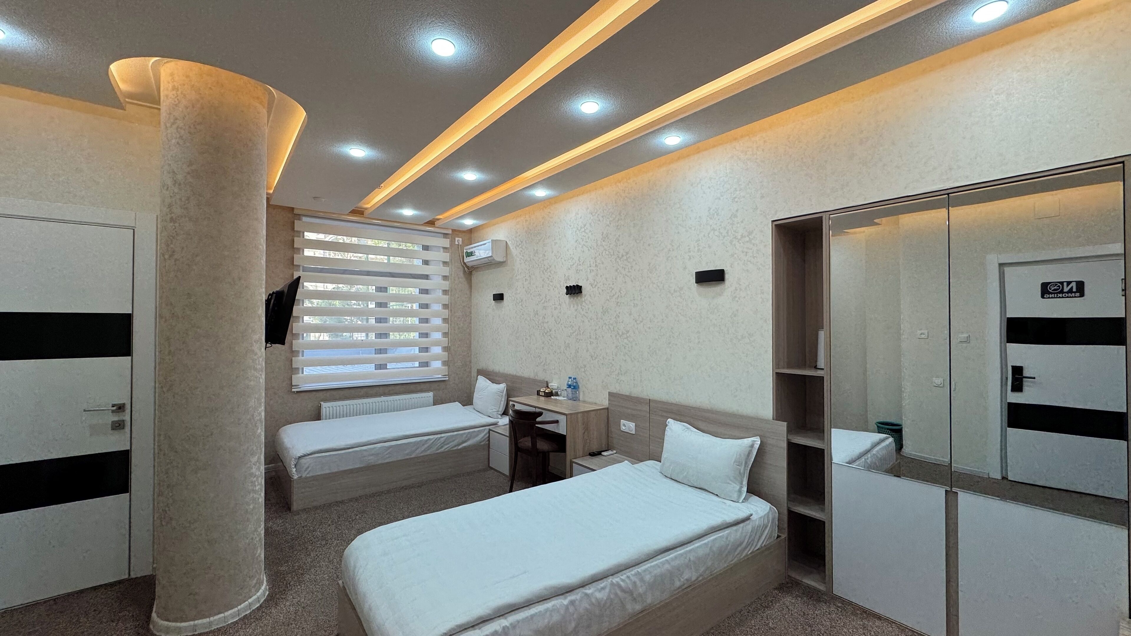 A hotel room with two single beds, a desk, a chair, a wardrobe, and a large pillar.