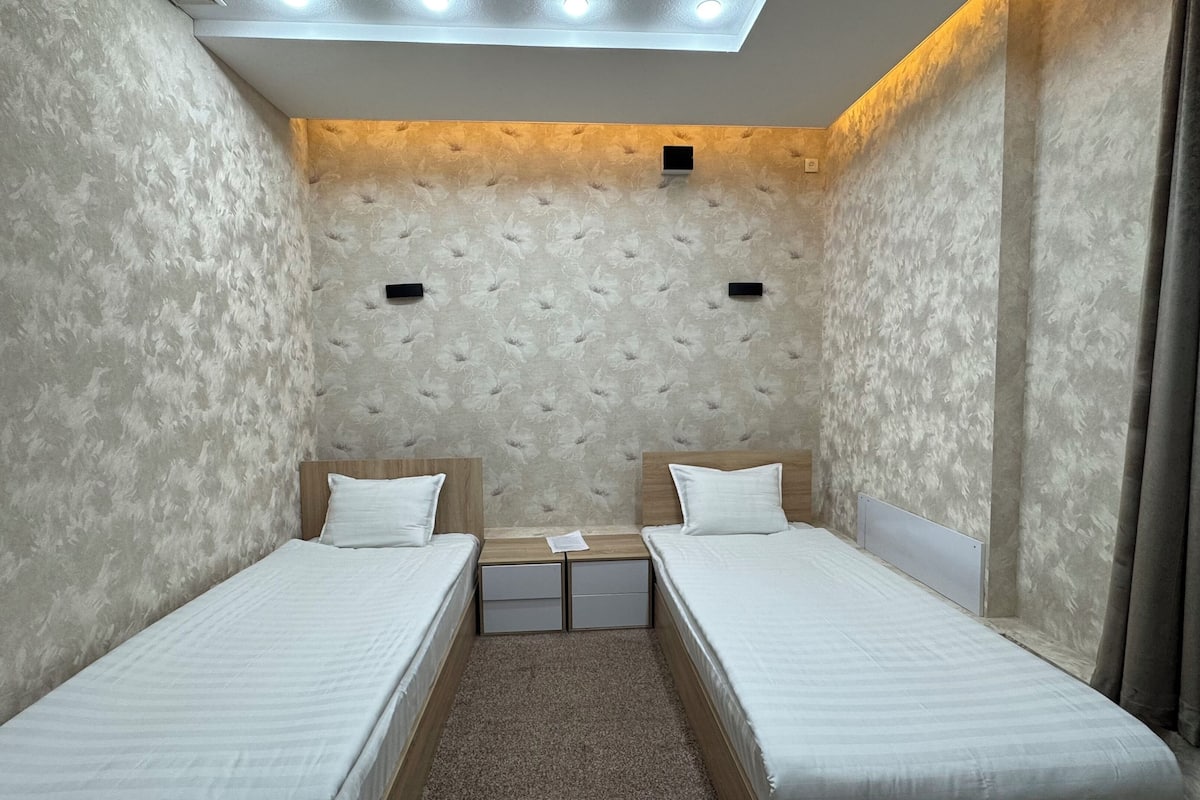 A small, cozy room with two single beds, a small table, and a ceiling light fixture.
