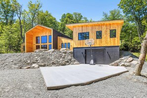 Una cabaña moderna de madera con aro de baloncesto, rodeada por un camino de grava y un montón de rocas.