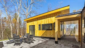 A modern wooden cabin with a covered patio area featuring outdoor seating and large windows. - Luxury Barn | Theater & Basketball Court (Winhall)