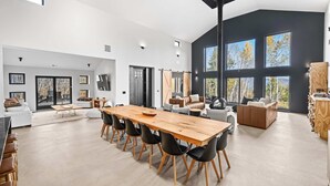 A spacious living area with a large wooden dining table, black chairs, and a sofa. There are multiple windows providing natural light and views of the outdoors. - Luxury Barn | Theater & Basketball Court (Winhall)
