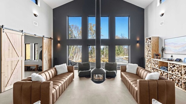A spacious living area with a large sectional sofa, a fireplace, and a view of the outdoors through large windows. - Luxury Barn | Theater & Basketball Court (Winhall)