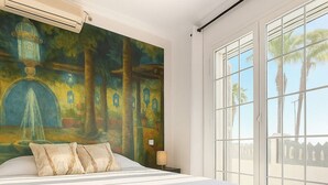 Ein Schlafzimmer mit einem großen Bett, einem Bild an der Wand und Blick auf Palmen durch eine Schiebetür.
