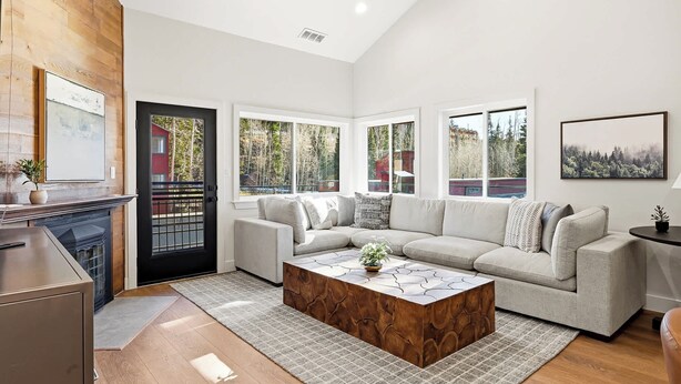 A modern living room with a sectional sofa, a wooden coffee table, and large windows offering a view of trees and a building. - Giant Steps 30 Ski in/Ski out (Brian Head)
