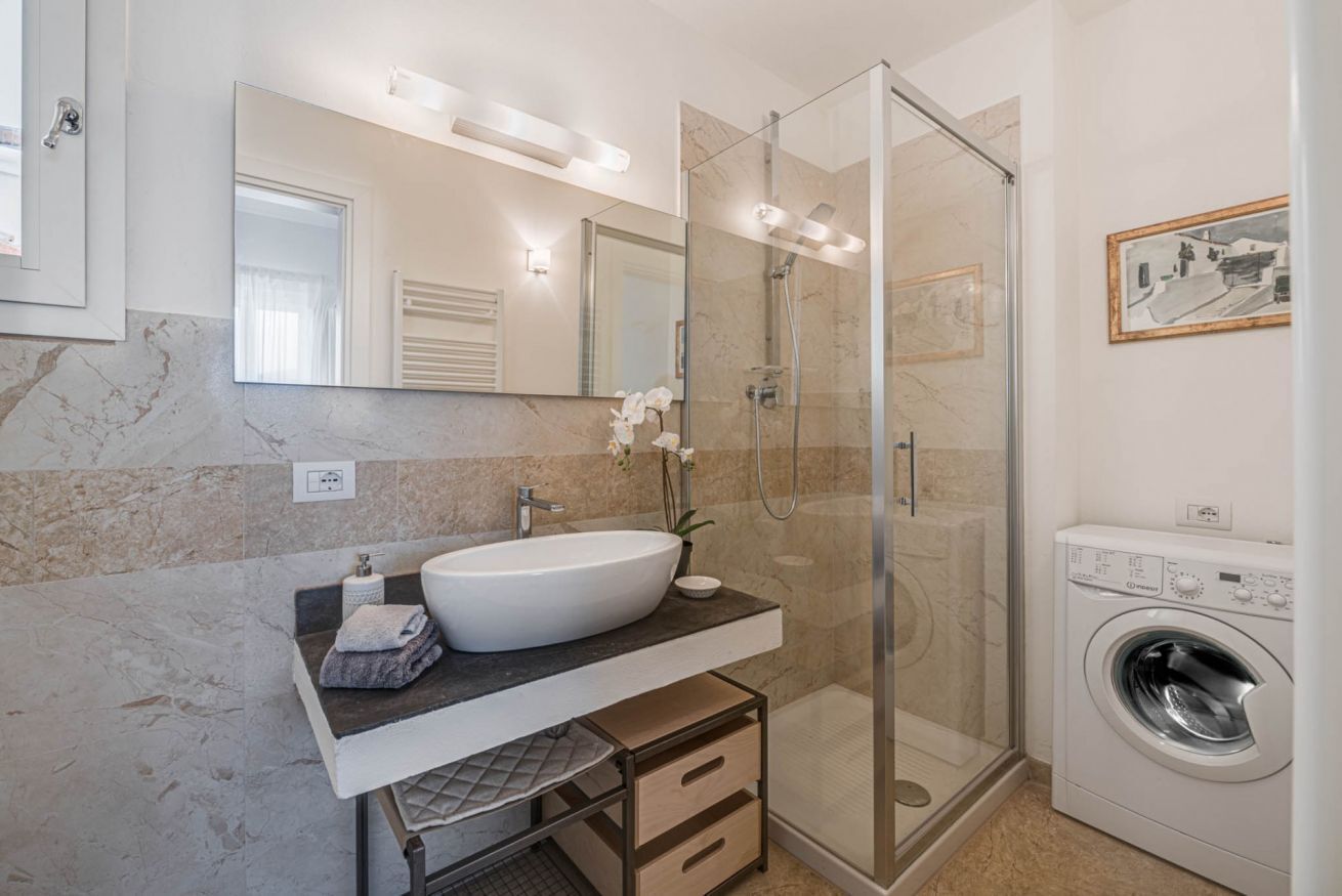 Un baño moderno con cabina de ducha de vidrio, lavamanos empotrado, lavadora y pared con azulejos de mármol.