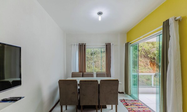A dining area with a table and chairs, a television on the wall, and a large window with curtains. - Apt 2 Bedrooms 200 m From the Beach Mao0203 (Ilhéus)