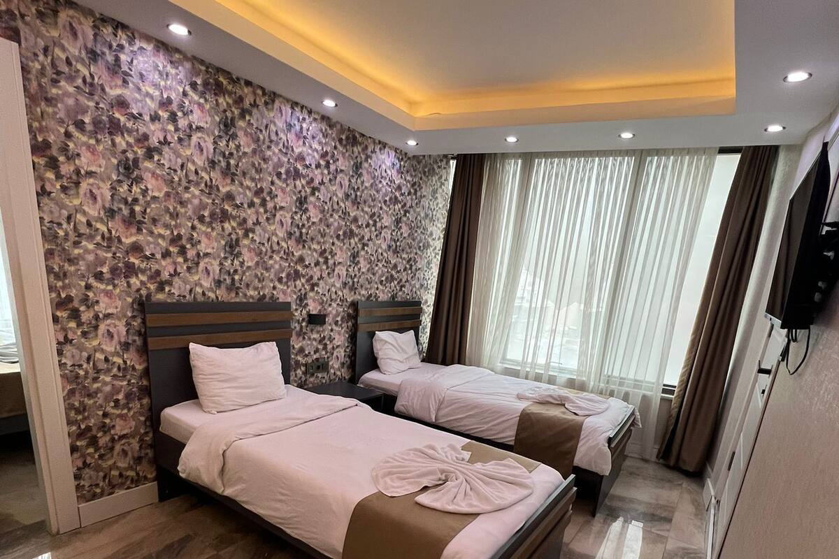 A hotel room with two beds, a stone-patterned wall, a large window with curtains, and a flat-screen TV.