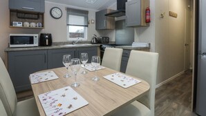 A compact kitchen with modern appliances, a dining table set with wine glasses, and a clock on the wall. - Woodland Way (Pentraeth)