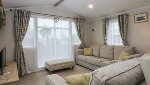 A modern living room with a sofa, ottoman, and a coffee table. There are curtains on the windows, and a picture frame on the wall. - Woodland Way (Pentraeth)