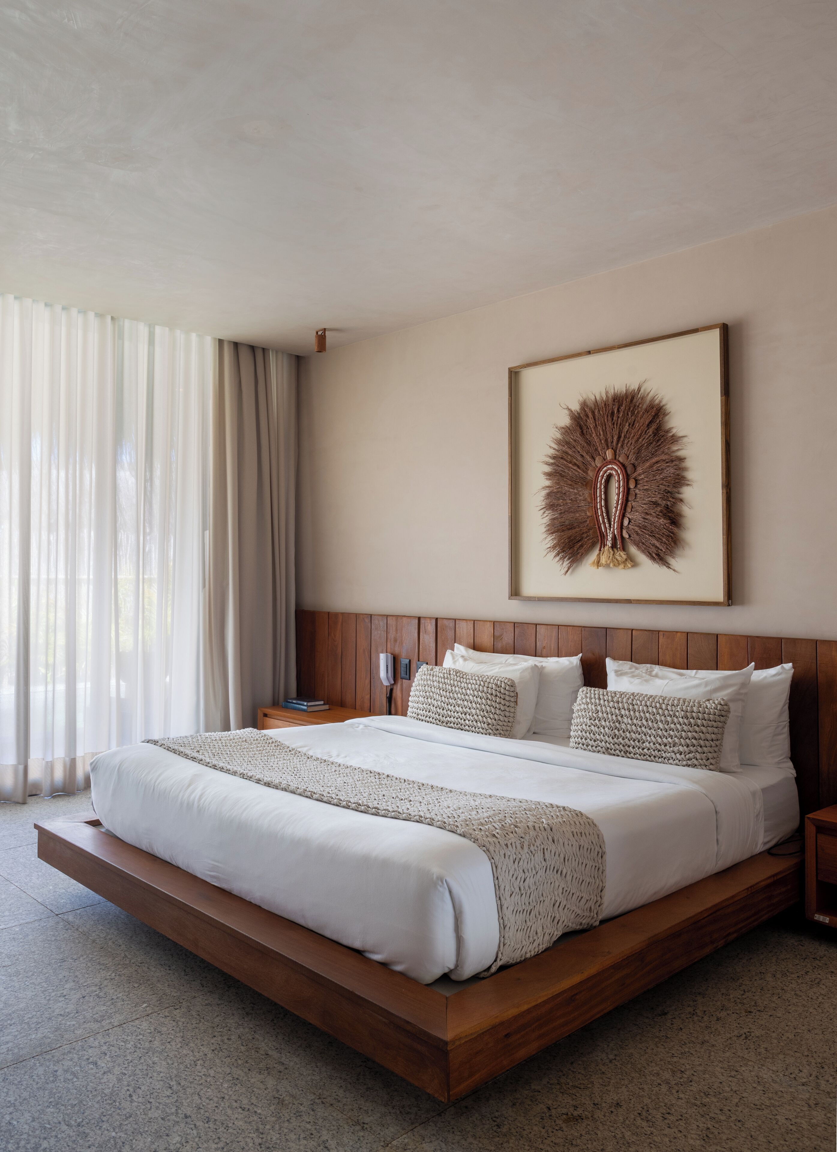 A hotel room with a large bed, wooden headboard, a framed artwork on the wall, and a nightstand with a lamp.