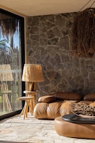 Quarto com parede de pedra, sofá de couro, luminária de chão trançada e vista através de uma grande janela.