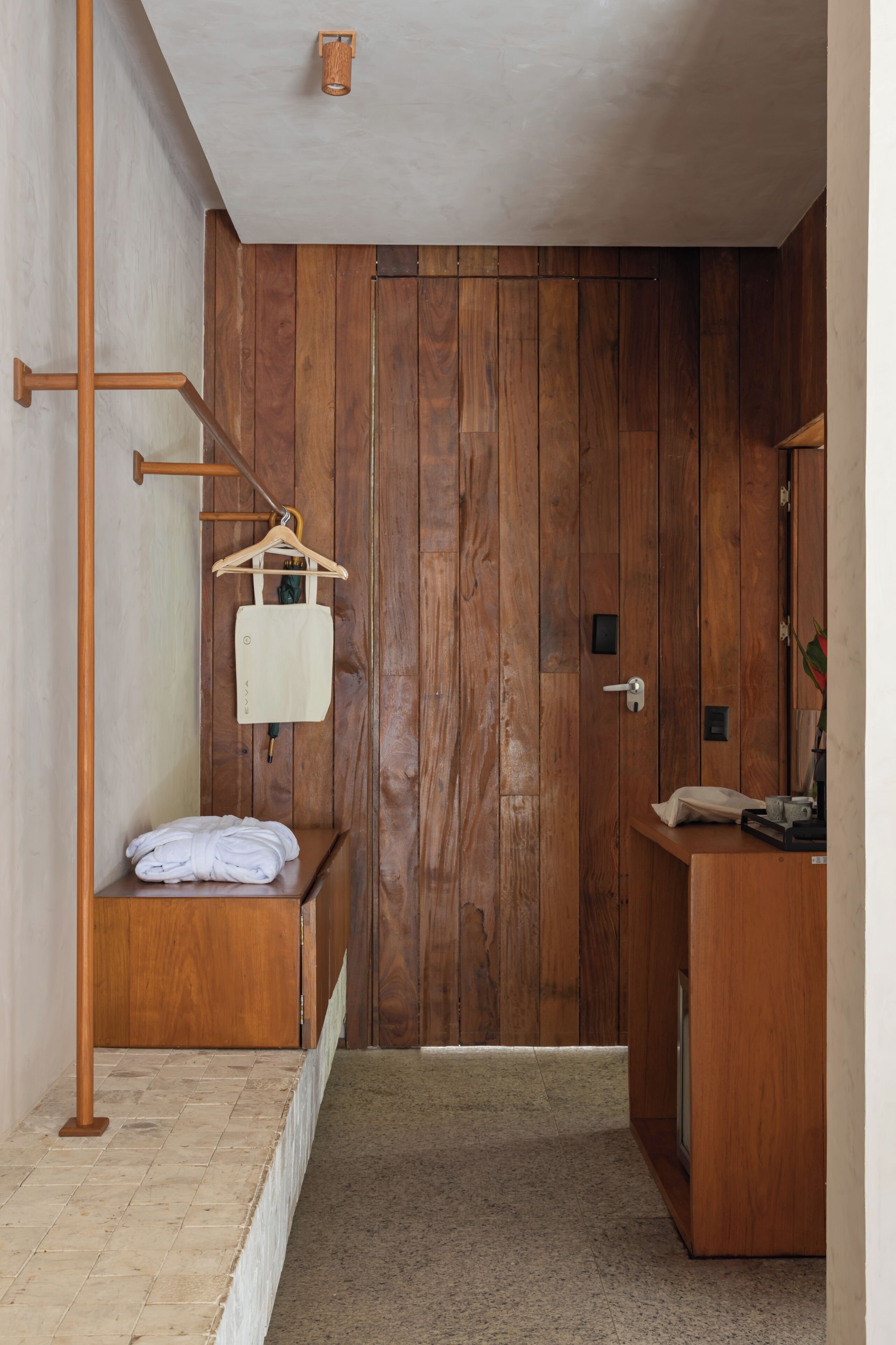 A wooden door with a towel rack and a hanging bag, a bench with a towel, and a small table with a cup and a tissue.