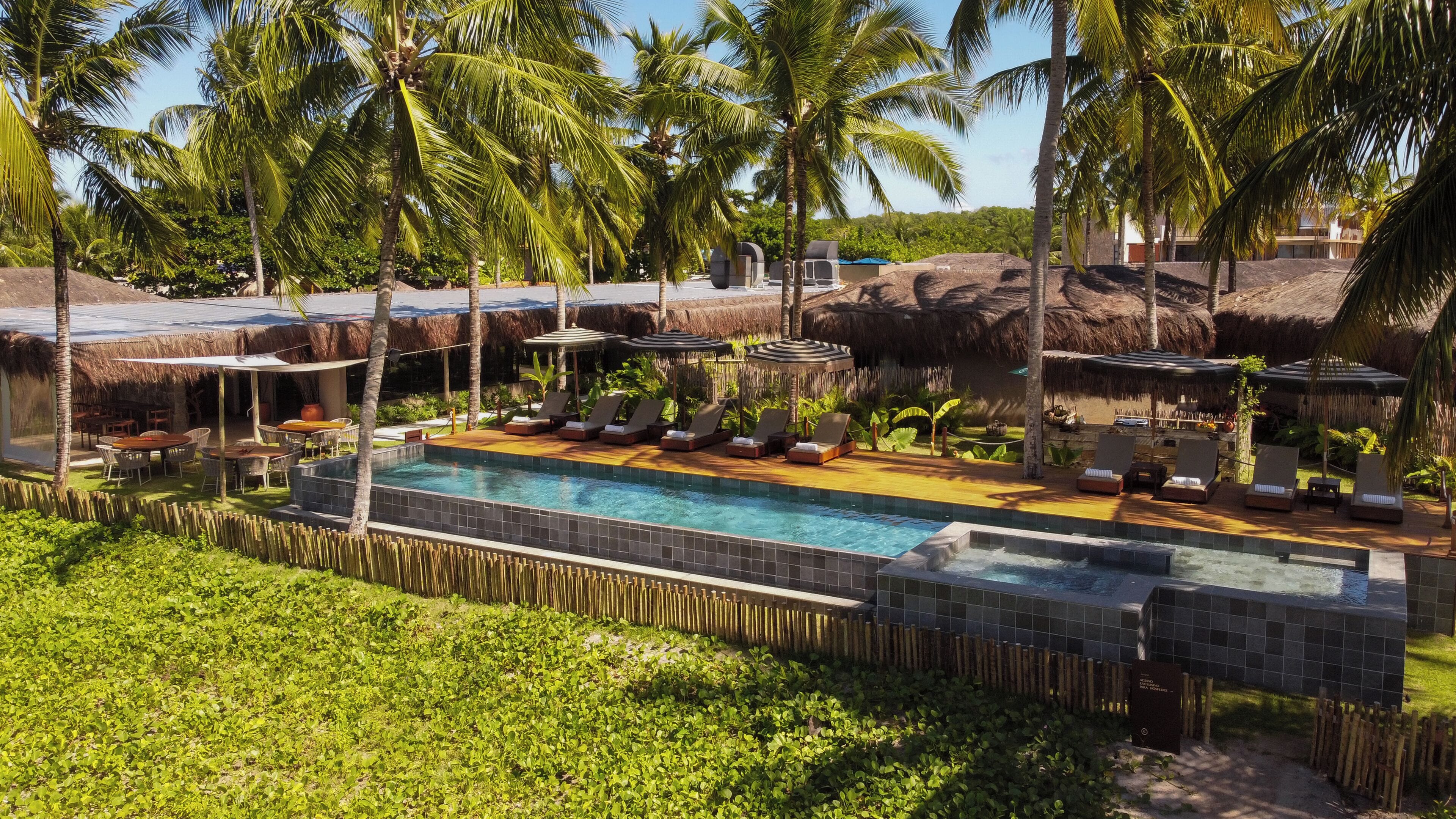 Un área de piscina con sillones reclinables y sombrillas, rodeada de palmeras y construcciones de paja.