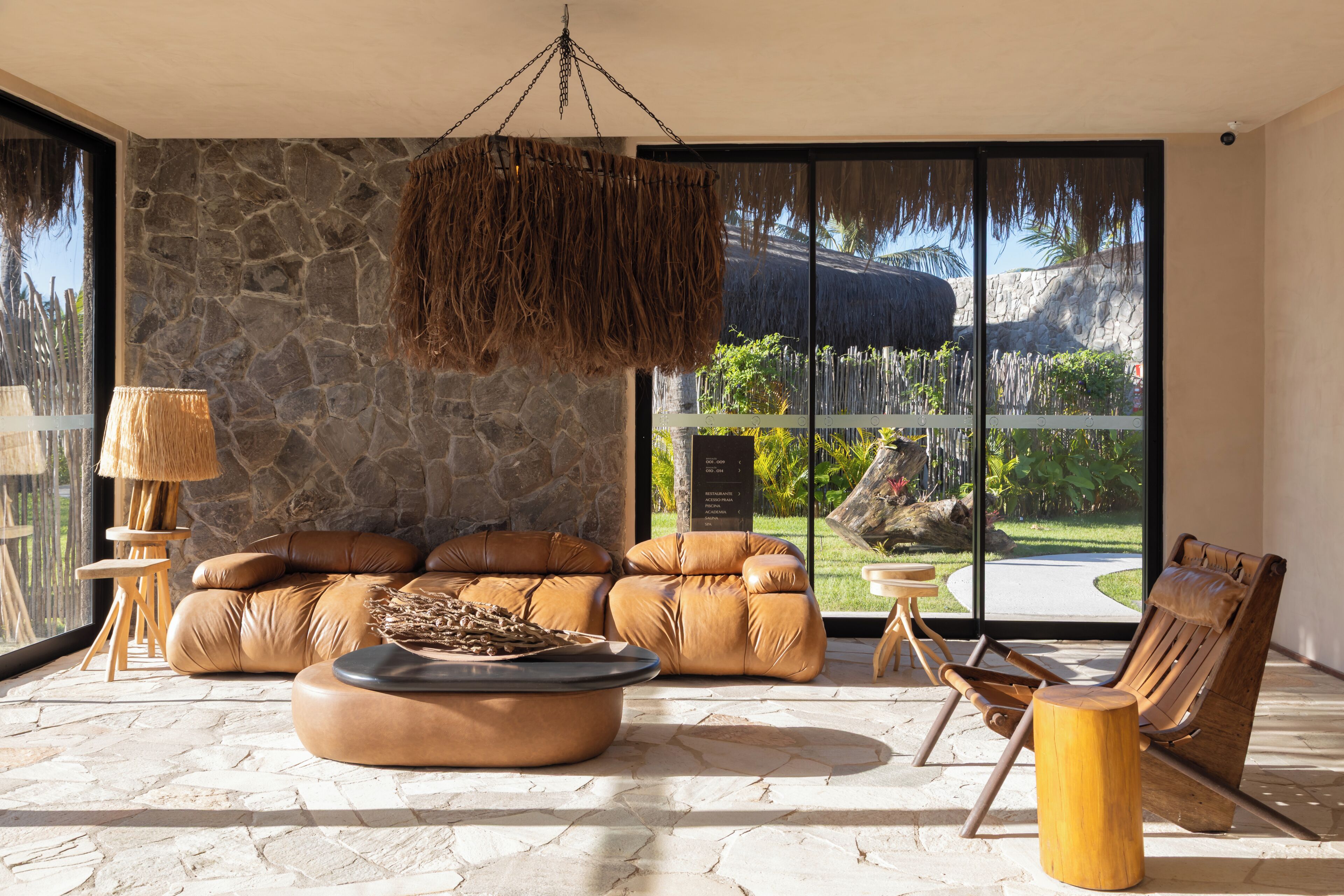 Una sala de estar moderna con pared de piedra, sofá de cuero y un ventanal con vistas a un paisaje tropical.