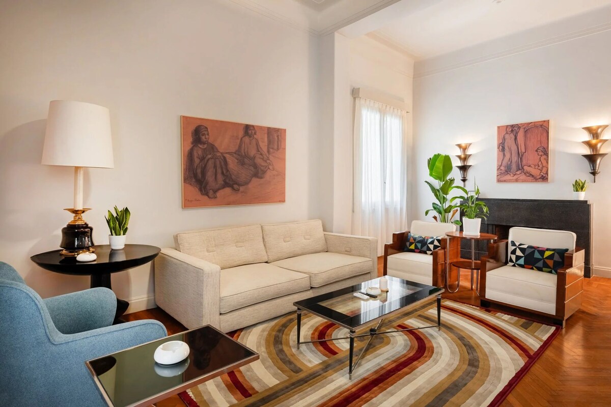 Una sala de estar con sofá, sillones, una mesa de centro, lámpara y obras de arte en las paredes.