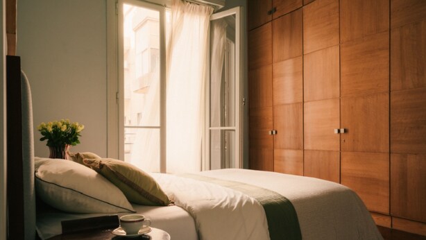 Un dormitorio con cama, almohadas, una mesita de noche con una taza y un armario de madera.