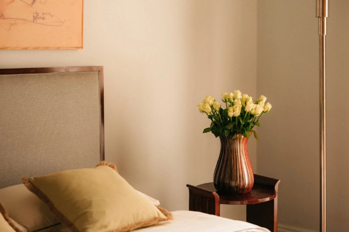 A bedroom with a bed, a bedside table with a vase of flowers, and a painting on the wall.