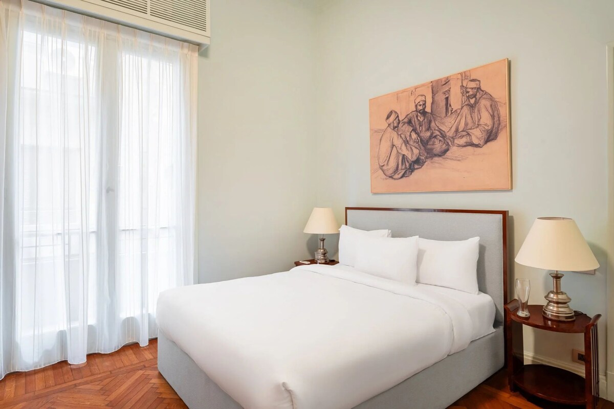 Una habitació d'hotel amb un llit gran, dues taules de nit amb làmpades, una finestra amb cortines translúcides i una obra d'art penjada a la paret.