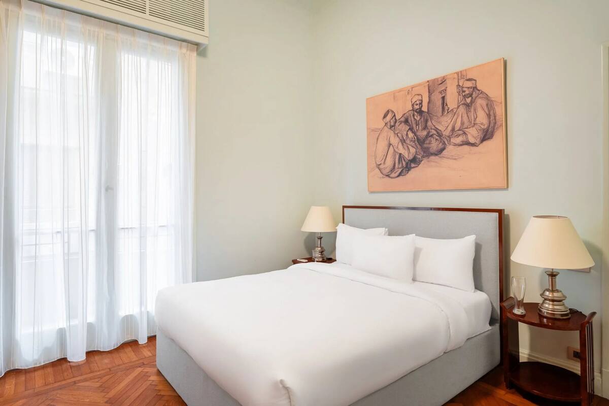 Habitación de hotel con una cama grande, dos mesitas de noche con lámparas, una ventana con cortinas traslúcidas y una obra de arte colgada en la pared.