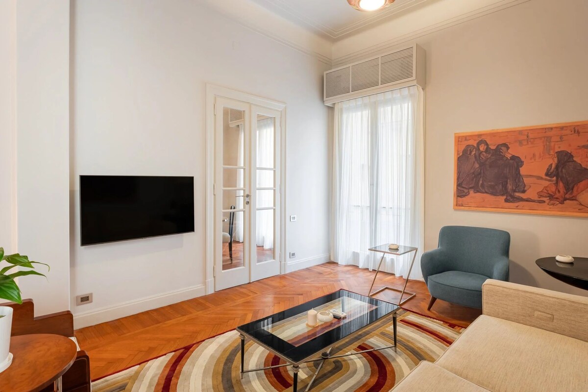 A modern living room with a flat-screen TV, a glass coffee table, a blue armchair, and a painting on the wall.