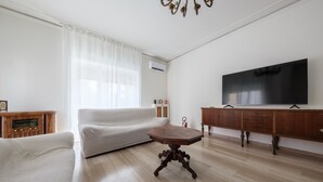 Ein Wohnzimmer mit einem weißen Sofa, einem Holztisch, einem Fernseher auf einer Holzkommode und einer weißen Heizkörper.