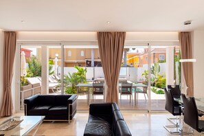 A modern living room with a glass door leading to an outdoor area, a black leather sofa, a glass coffee table, and a TV. - Villa Deluxe Suites Maspalomas (Maspalomas)