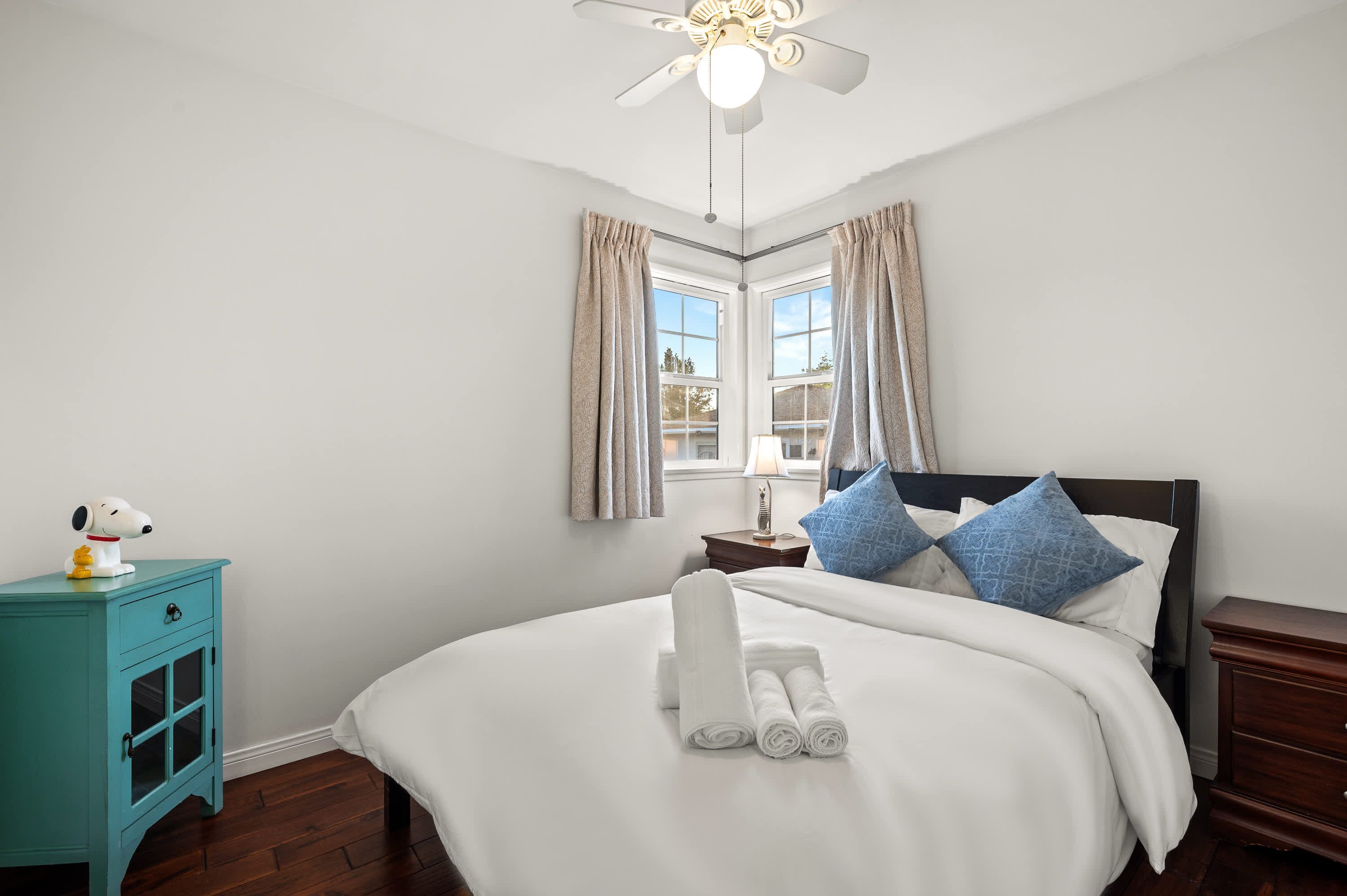 A neatly made bed with rolled towels, a nightstand with a lamp, a ceiling fan, and a window with curtains.