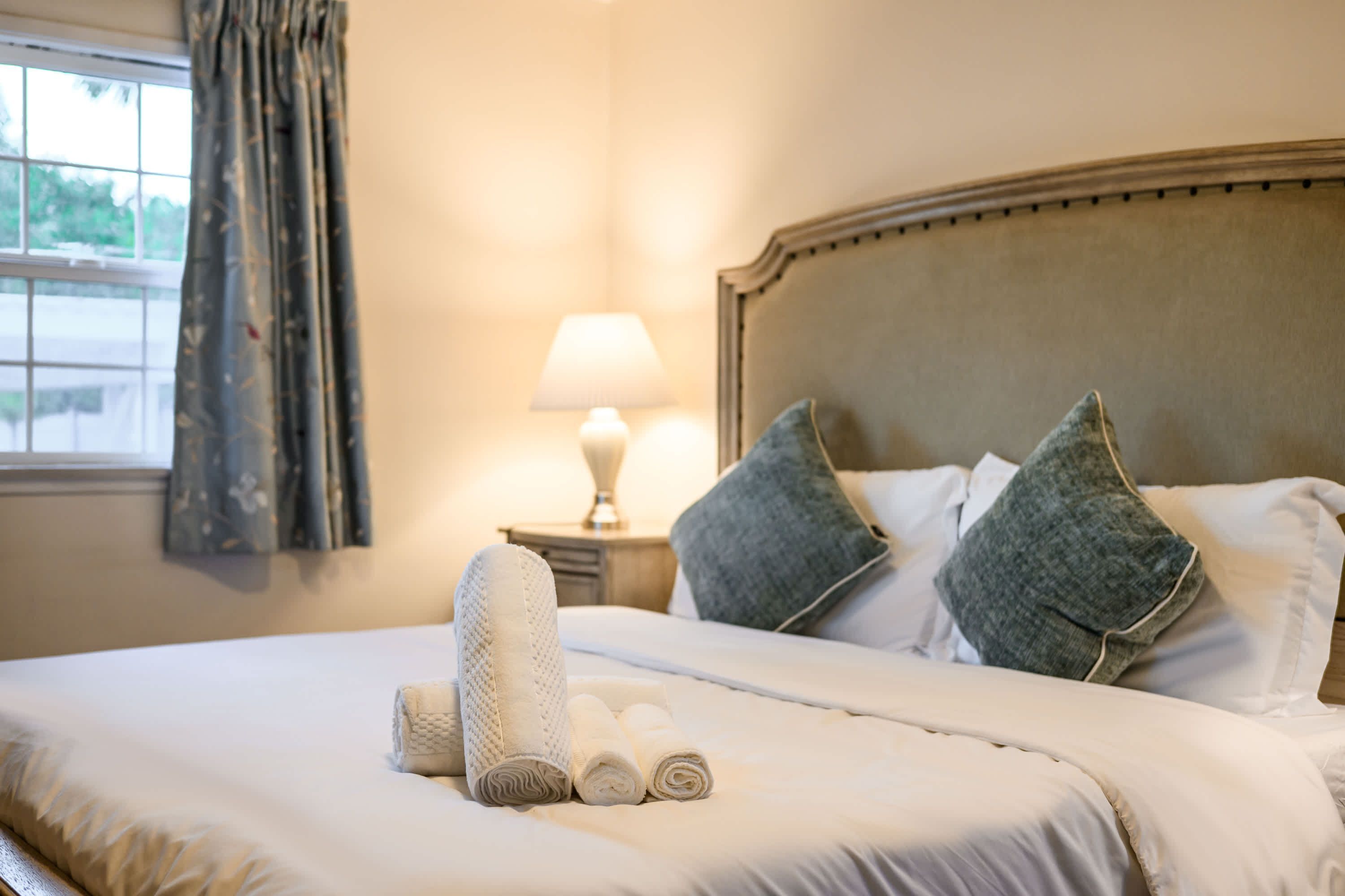 A neatly made bed with rolled towels and pillows, a bedside lamp, and a window with curtains.