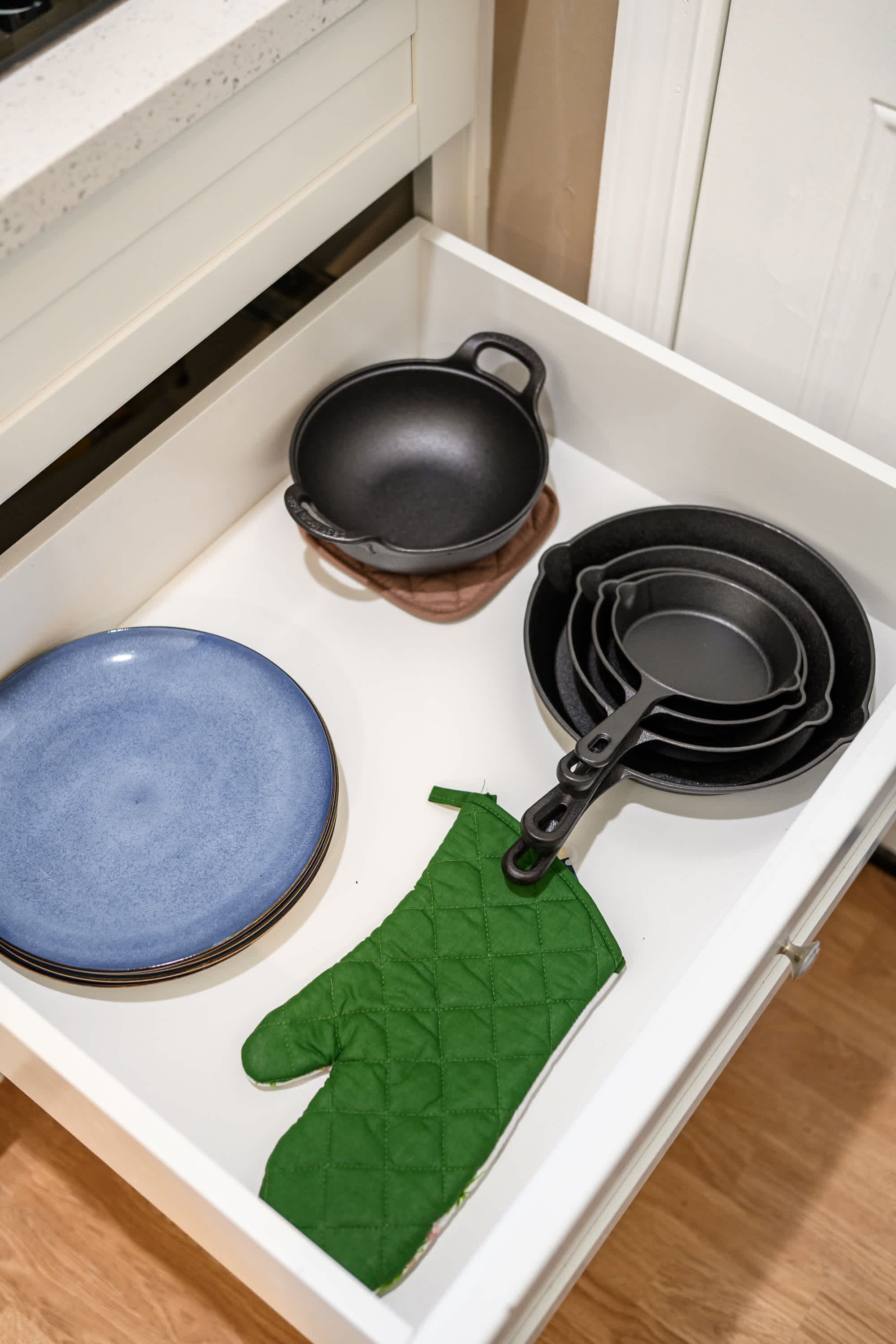 A kitchen drawer containing a blue plate, a black frying pan, a green oven mitt, and a black saucepan.