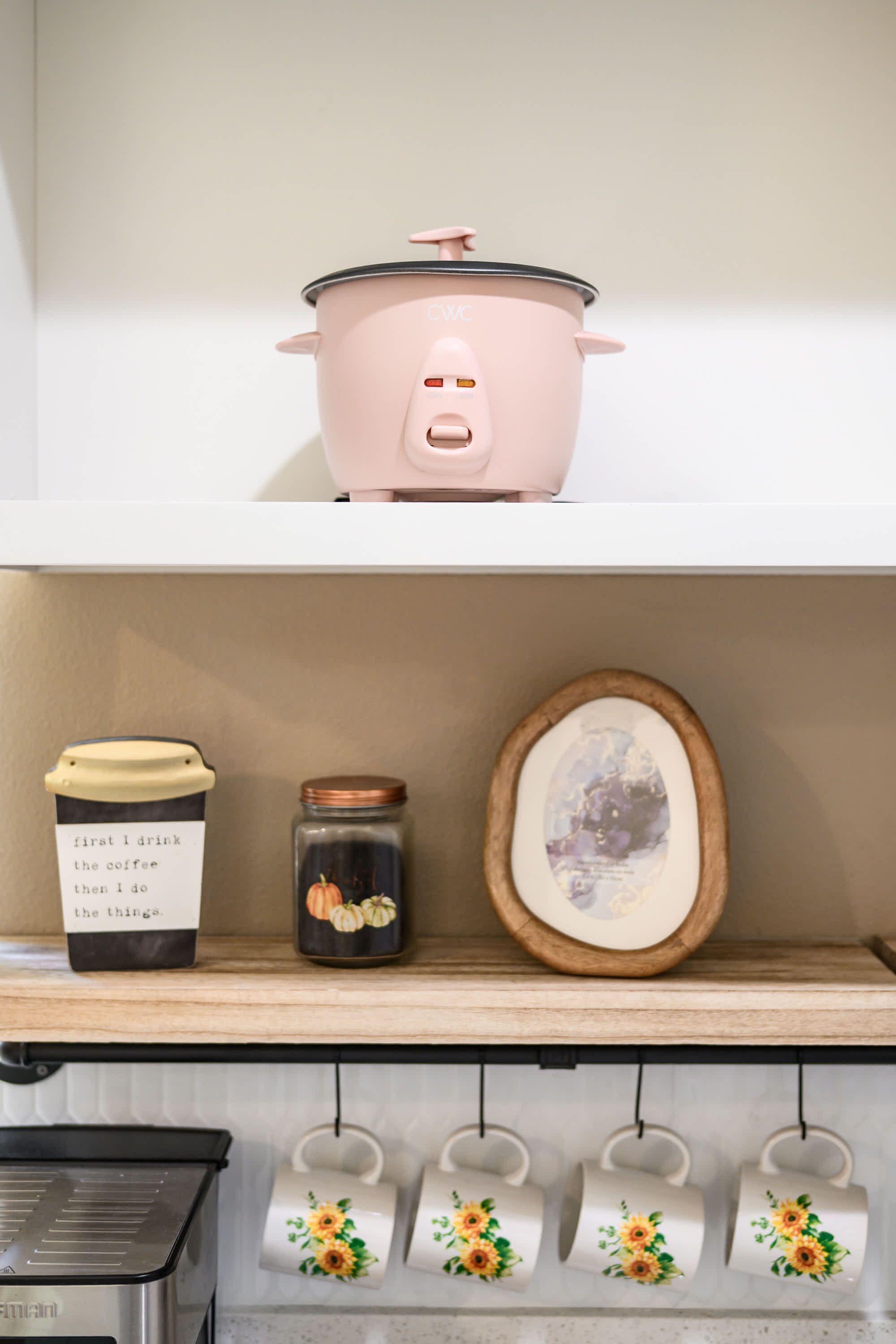 A kitchen with a pink slow cooker on the shelf, a coffee maker, a jar with a label, a framed picture, and several mugs with floral designs.