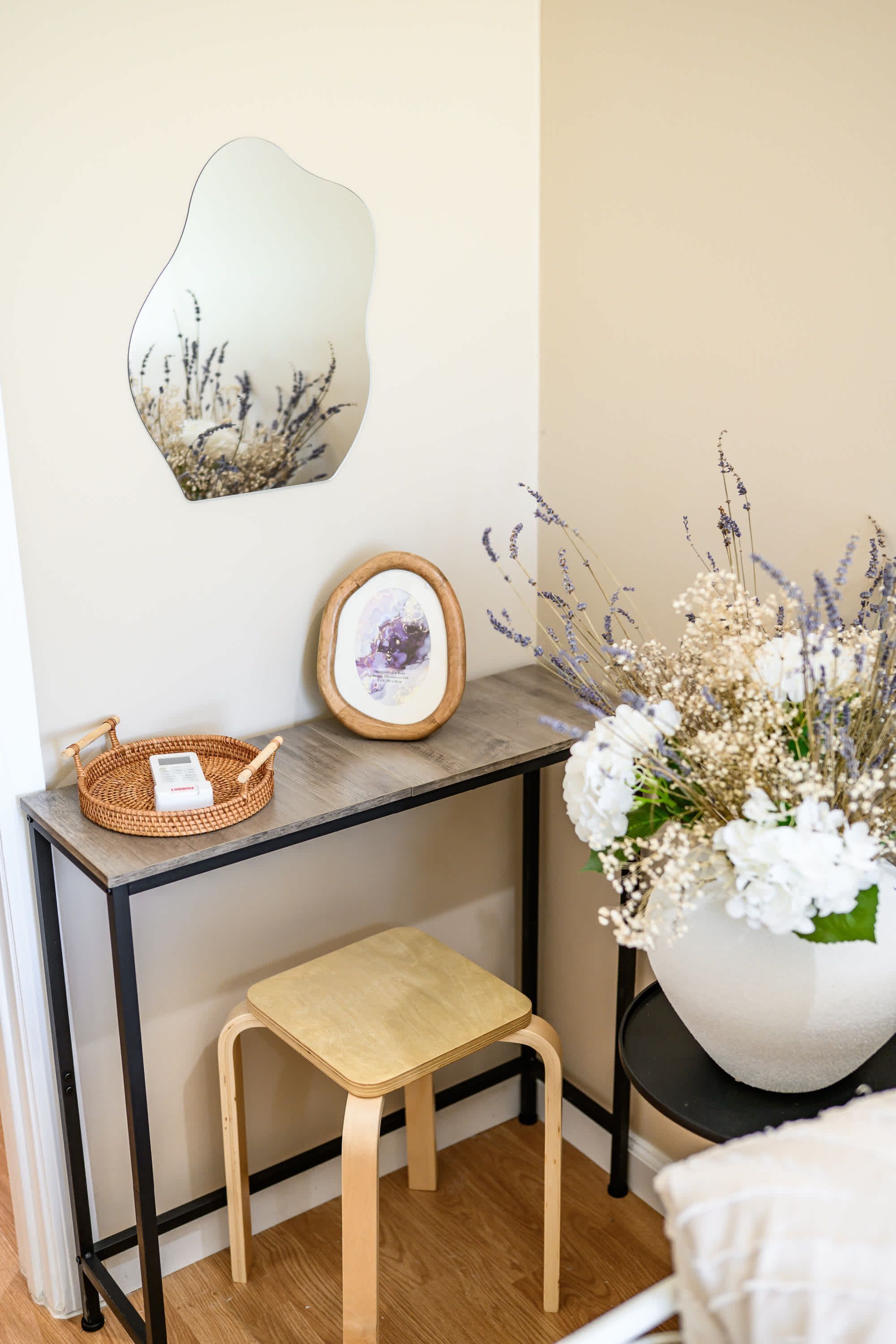 Eine Ecke eines Raumes mit einem hölzernen Hocker, einem gewebten Korb, einem gerahmten Bild und einer Vase mit Blumen.