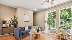 Ein Wohnzimmer mit Sofa, Couchtisch und Sesseln, einem Esstisch mit Stühlen, einem Deckenventilator und großen Fenstern mit Blick auf Grünflächen.