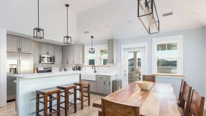 Cozinha moderna com armários cinza, ilha de madeira com banquetas, mesa de jantar grande de madeira e iluminação pendente.
