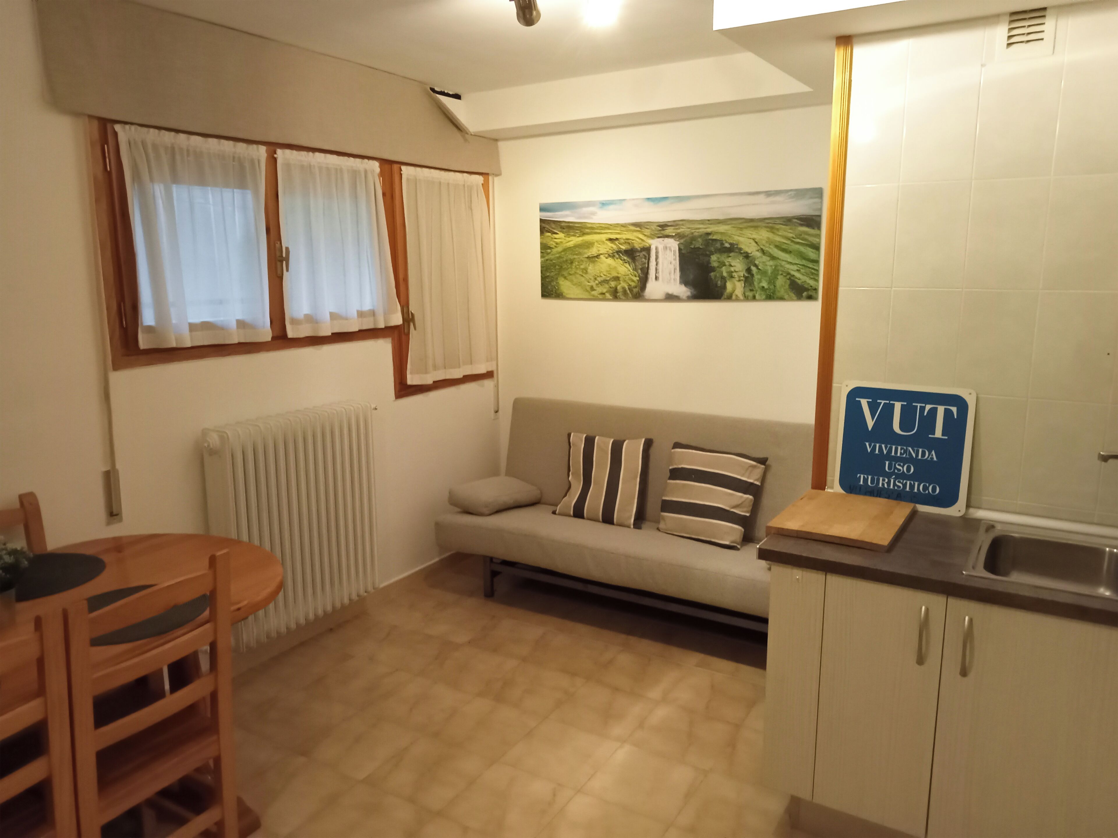 A compact kitchen with a dining area, a sofa with striped cushions, a radiator, and a painting of a waterfall on the wall.