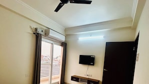 Quarto de hotel com cama, ventoinha no teto, TV fixada na parede e janela com cortinas.