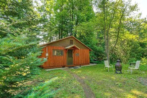 A small wooden cabin with a door and windows, surrounded by lush greenery and a path leading to it. - Cozy 'rivershire Retreat Cabin' Near Traverse City (Traverse City)