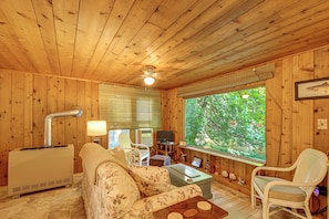 A wooden cabin interior with a sofa, a table, a chair, a television, and a window with a view of greenery. - Cozy 'rivershire Retreat Cabin' Near Traverse City (Traverse City)