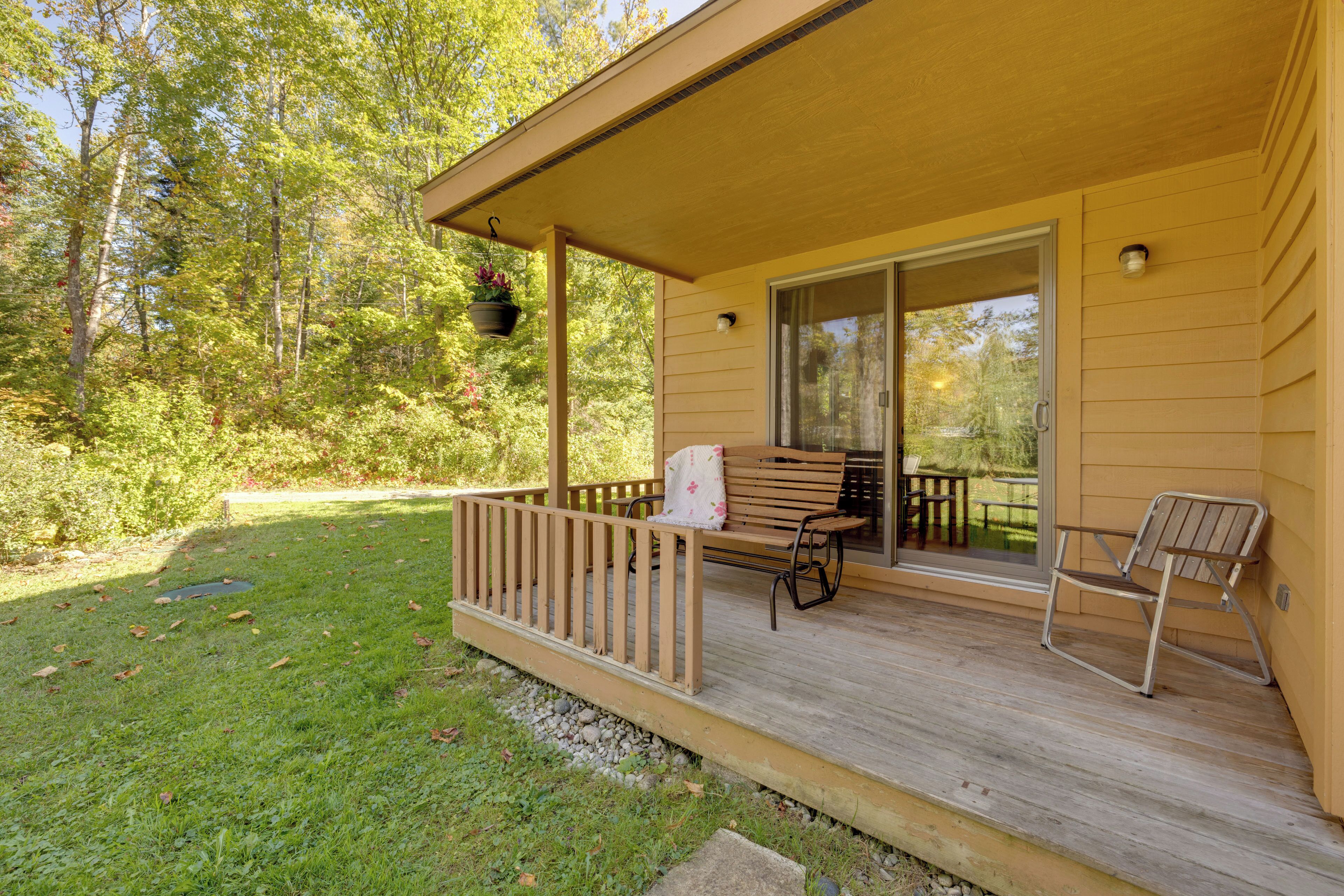 Une terrasse en bois avec un banc et une chaise, un pot de fleurs suspendu et une porte-fenêtre coulissante donnant sur un patio.