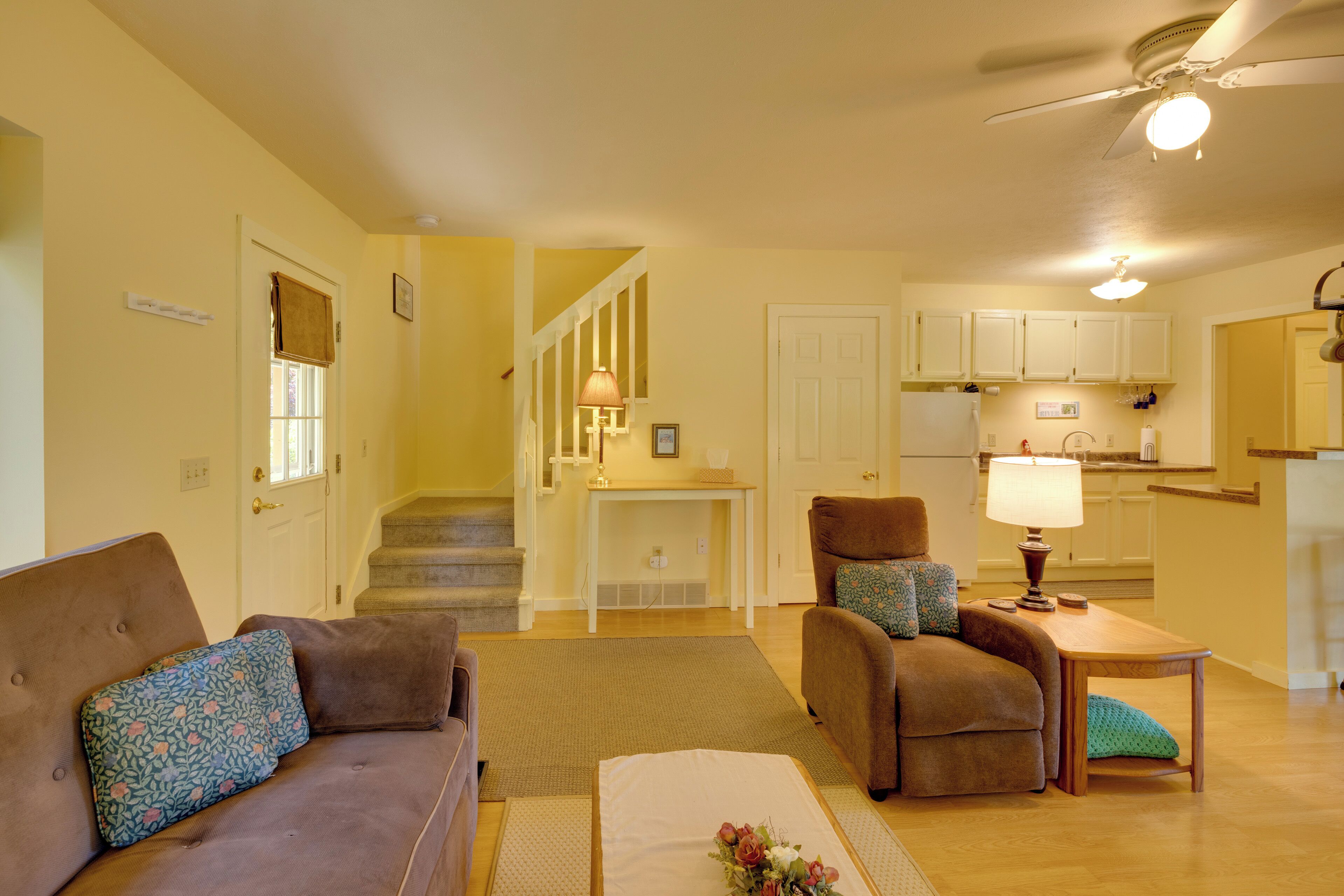 A living room with a brown sofa, a chair, a small table with a lamp, and a staircase leading to a kitchen.