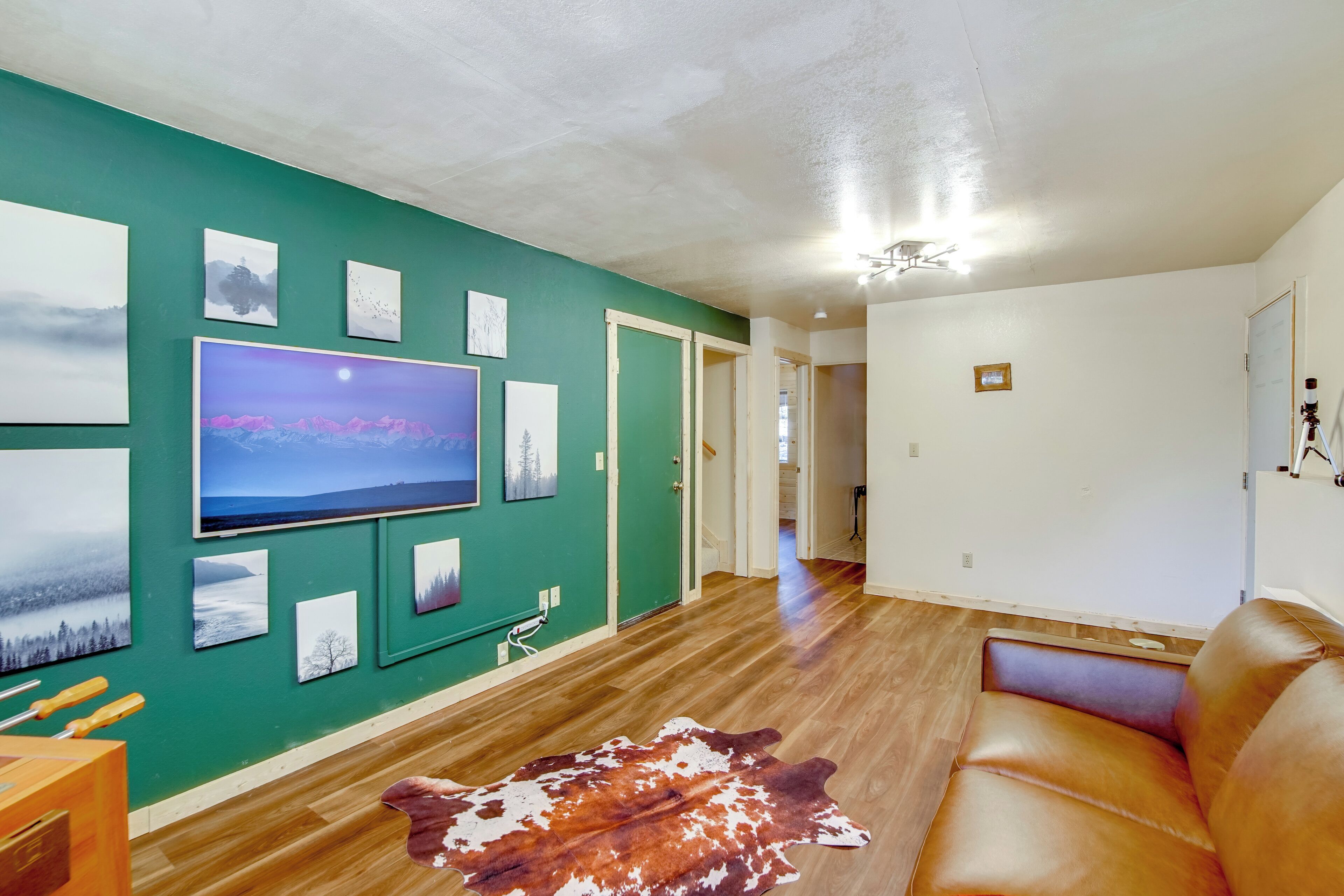 A living room with a large flat-screen TV, a leather sofa, a wooden floor, and a green accent wall with framed pictures.
