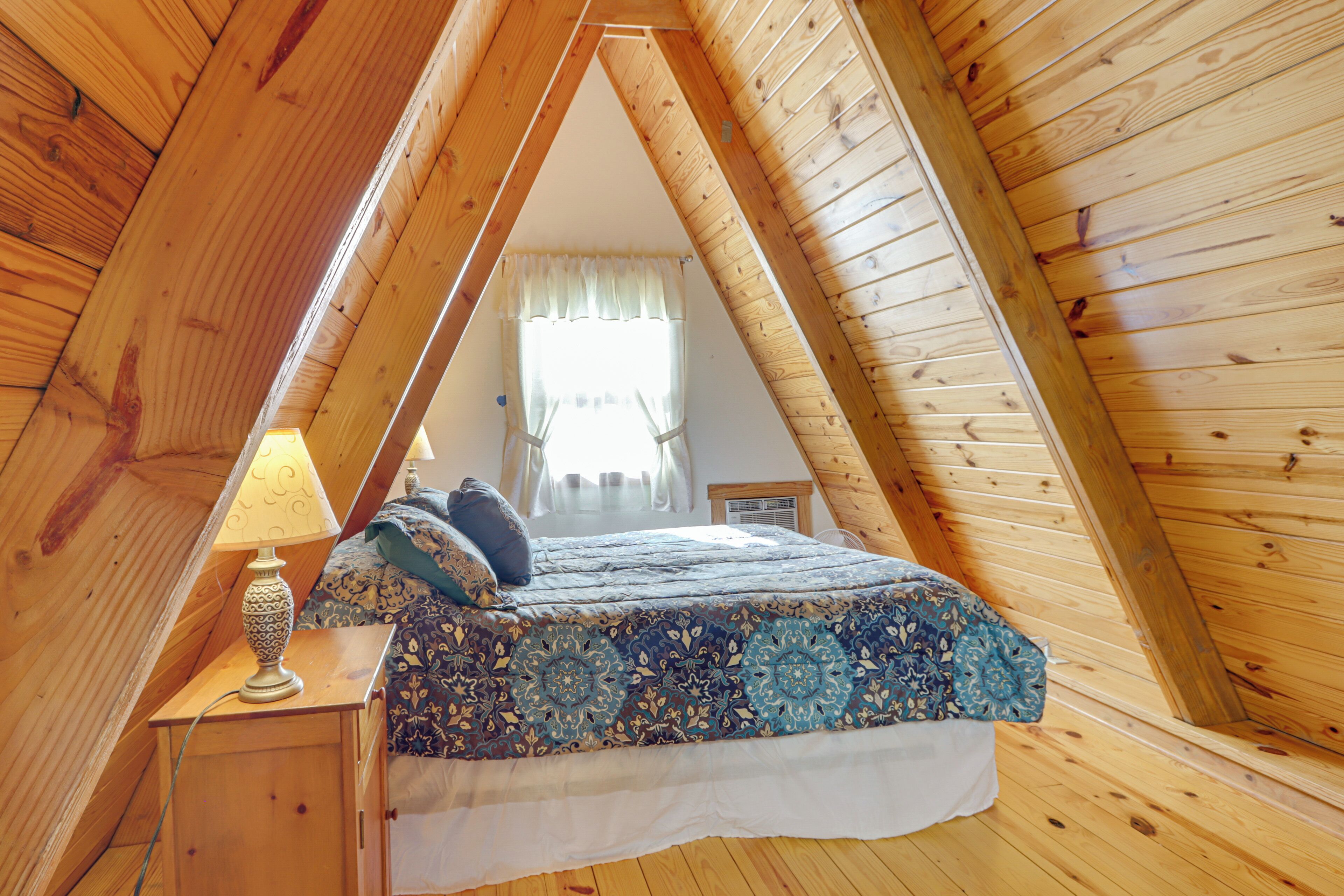 Une chambre avec un plafond en bois et un toit en pente, un lit avec une literie à motifs, une table de chevet avec une lampe, et une fenêtre avec des rideaux.