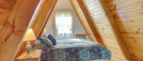 A bedroom with a wooden ceiling and sloped roof, a bed with patterned bedding, a bedside table with a lamp, and a window with curtains.