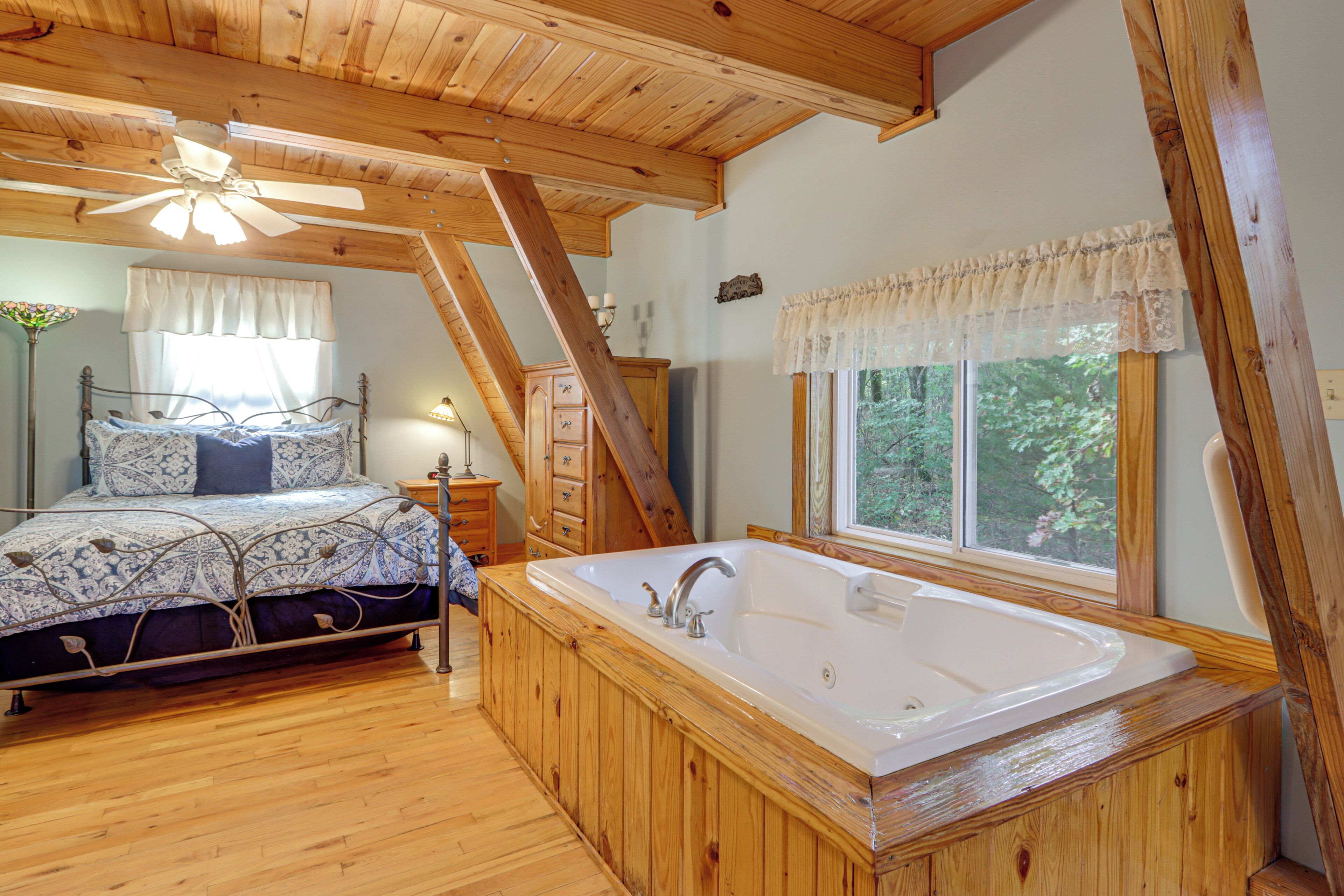 Ein Schlafzimmer mit Holzboden, einem großen Fenster, einem Bett mit gemusterten Betttextilien, einer Holzbadewanne und einem Nachttisch mit Lampe.